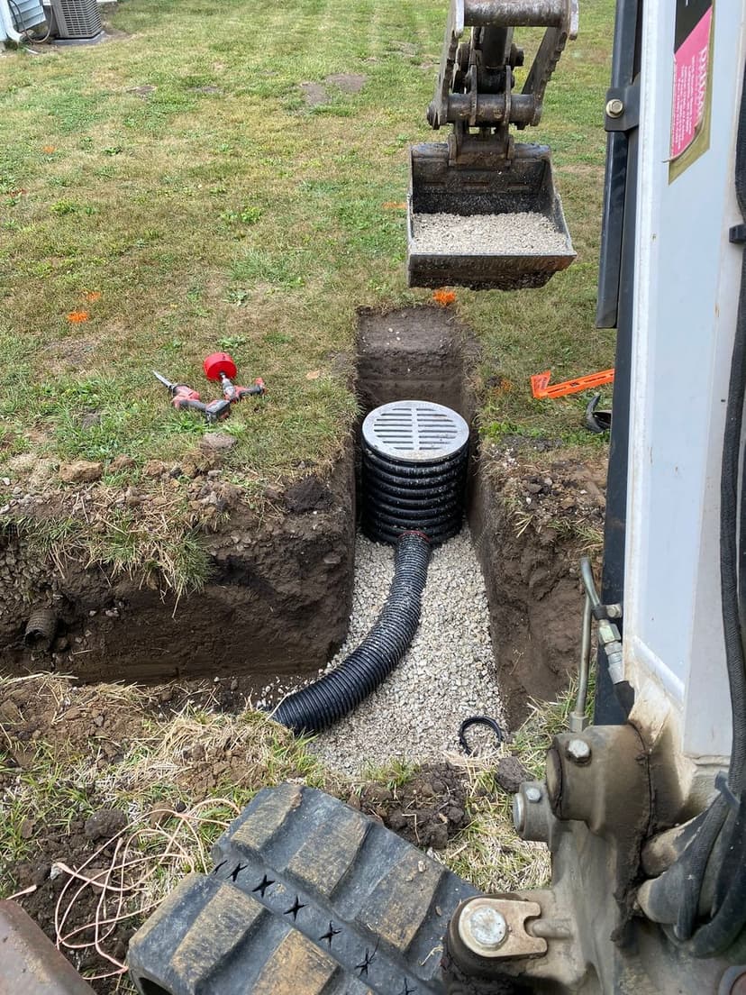 Excavation for drainage installation with gravel, pipe, and heavy machinery in a yard.