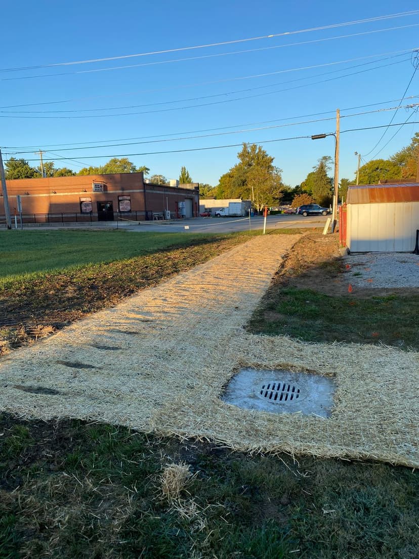 Pathway covered with straw leading to a drainage area near buildings and electric poles.