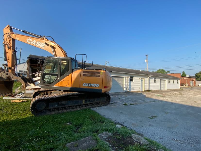 Case CX210D excavator parked beside a building on a clear day with blue sky.