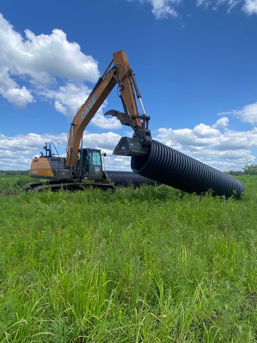 Excavator lifting large pipe in a grassy field under a blue sky with clouds.